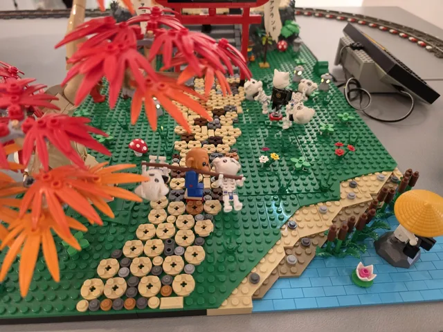 Eine japanisch gestaltete LEGO-Landschaft mit roten und orangen Ahornbäumen, Trittsteinen, einem roten Torii und kleinen weißen Wesen auf der Wiese